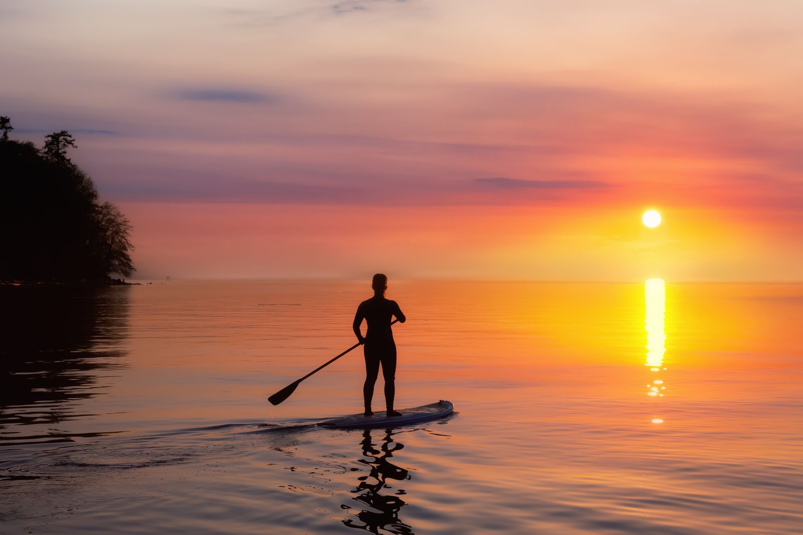 Silhouette of a surfer with a paddle standing on a paddleboard, gliding over calm waters against a stunning sunset, reflecting the serene and beautiful ambiance of paddleboarding.