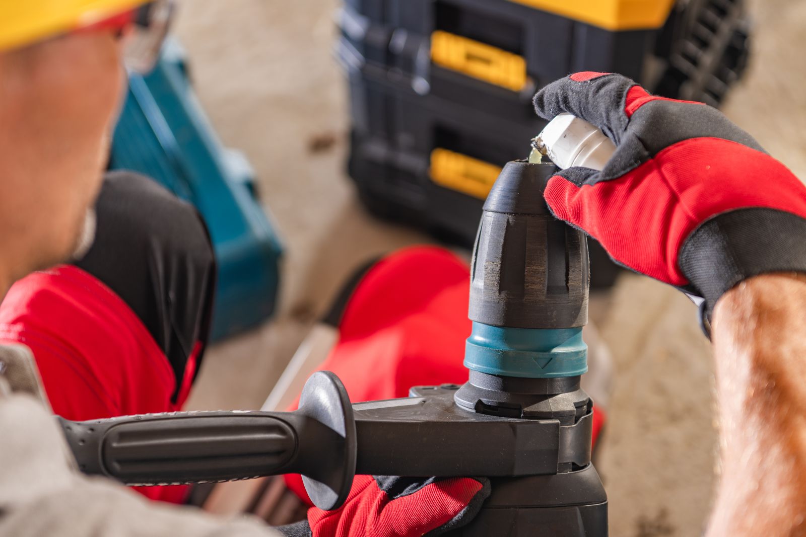 Close-up of a worker in a red shirt and black gloves using a professional blue power drill on a construction site, with yellow and blue toolboxes in the background.