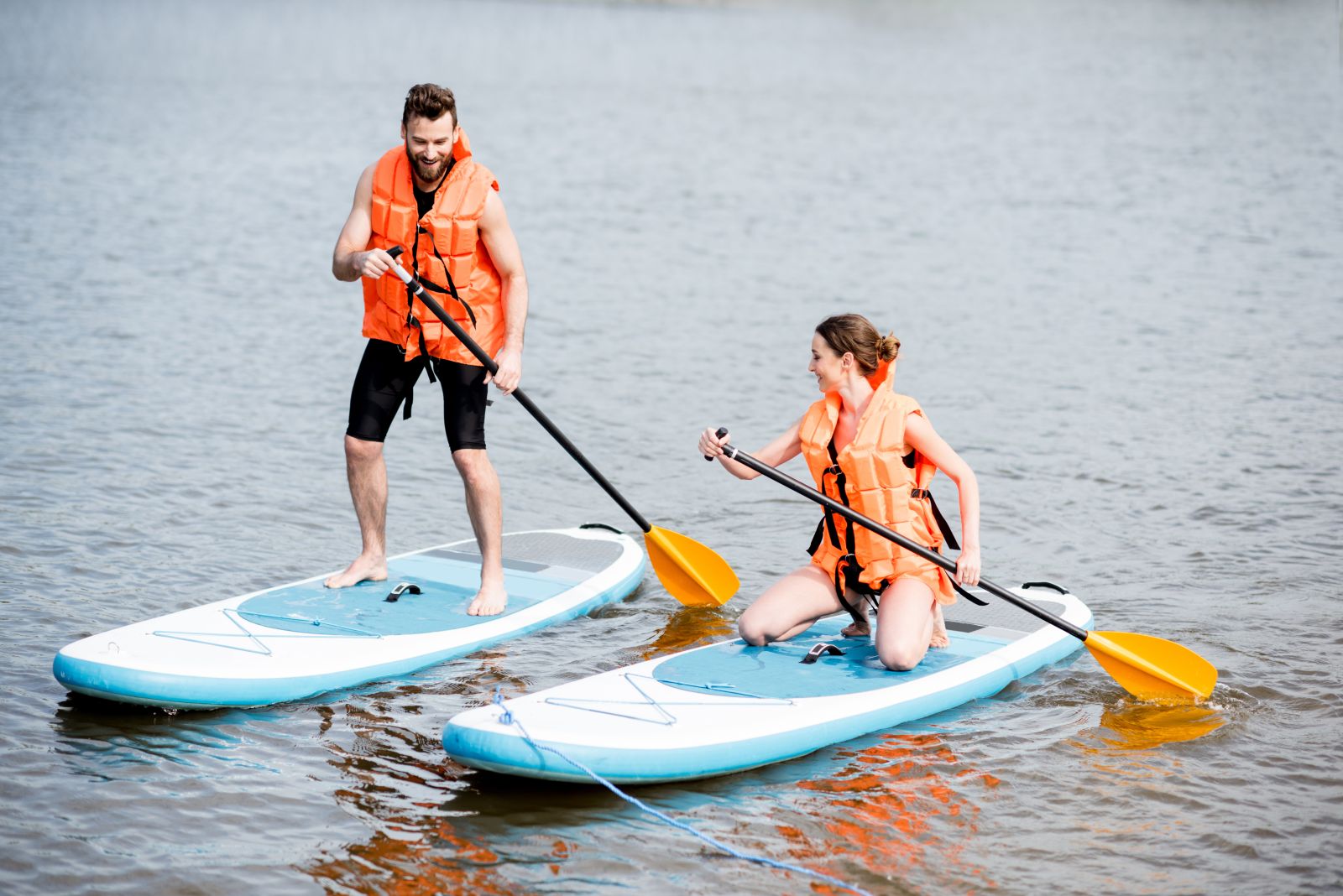 Two people joyfully engage in paddle boarding on a calm lake, both standing on blue paddleboards and wearing orange life jackets, showcasing an active and fun outdoor water activity.