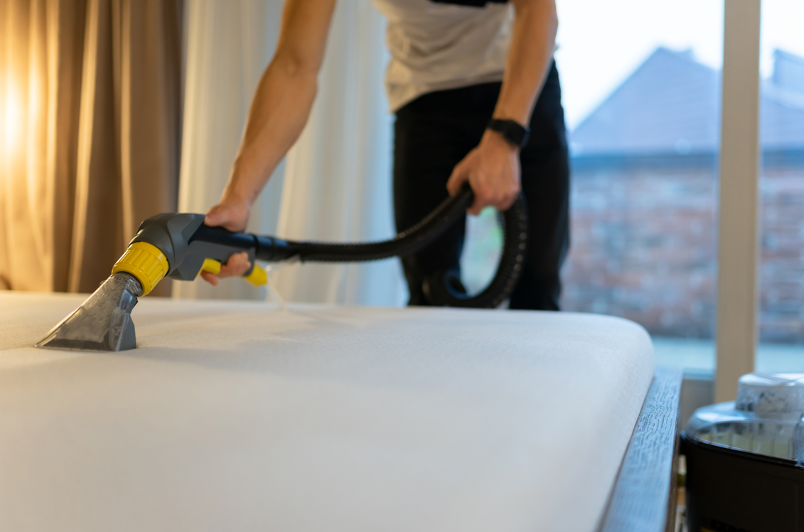 Close-up of a handheld steam cleaner being used on a white foam mattress.