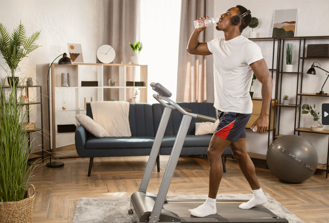 A man in a white t-shirt and red and blue shorts hydrating while using a treadmill at home, surrounded by stylish home decor and listening to music through headphones.