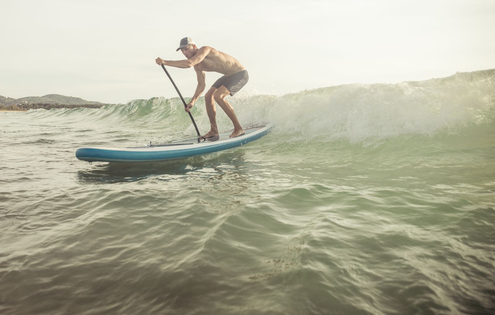An active male surfer with a paddle balances on a blue paddleboard, skillfully navigating a wave, demonstrating a dynamic moment of water sports adventure.
