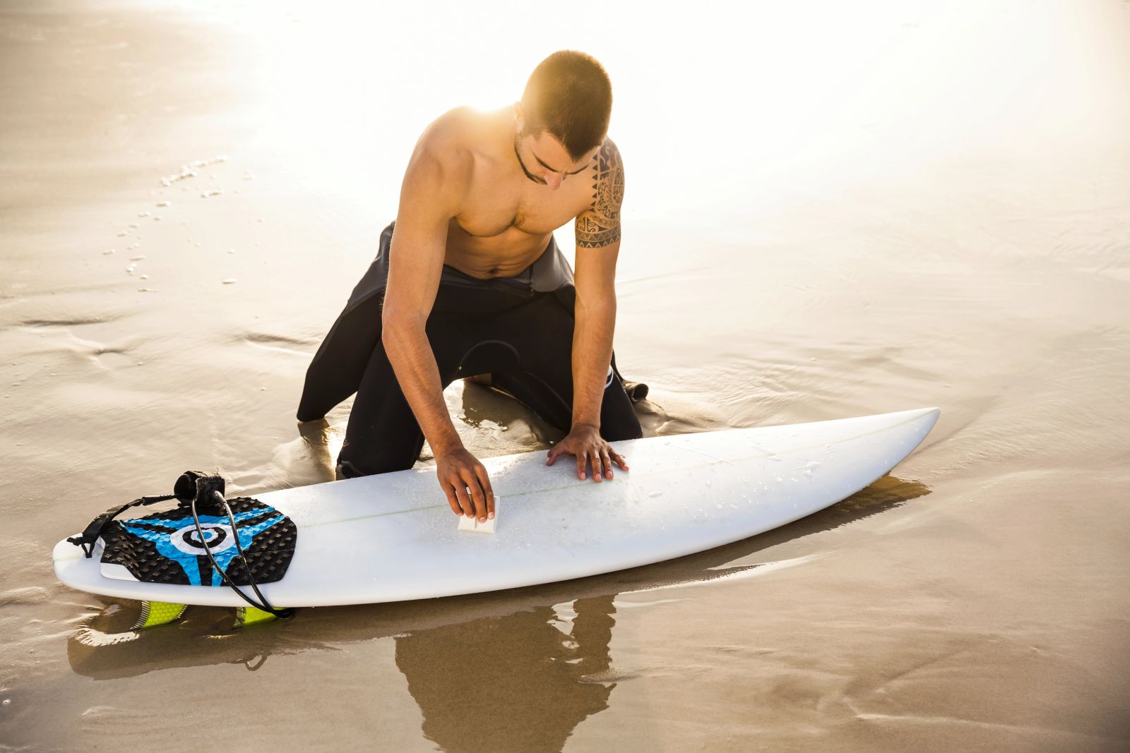 A surfer kneels on the beach, applying wax to his surfboard. This image illustrates one answer to “Why do surfers wax their surfboards?” with that answer being: to ensure better grip and control.